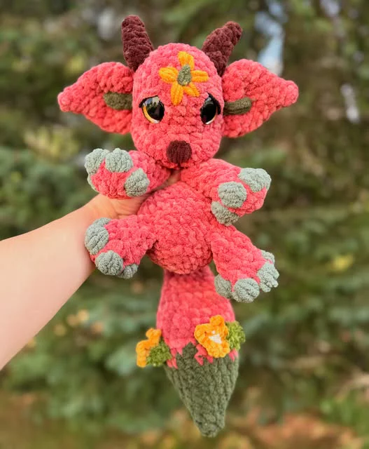 A red crocheted strawberry with safety eyes, a stitched smile, green leaf arms and a white flower with a yellow centre