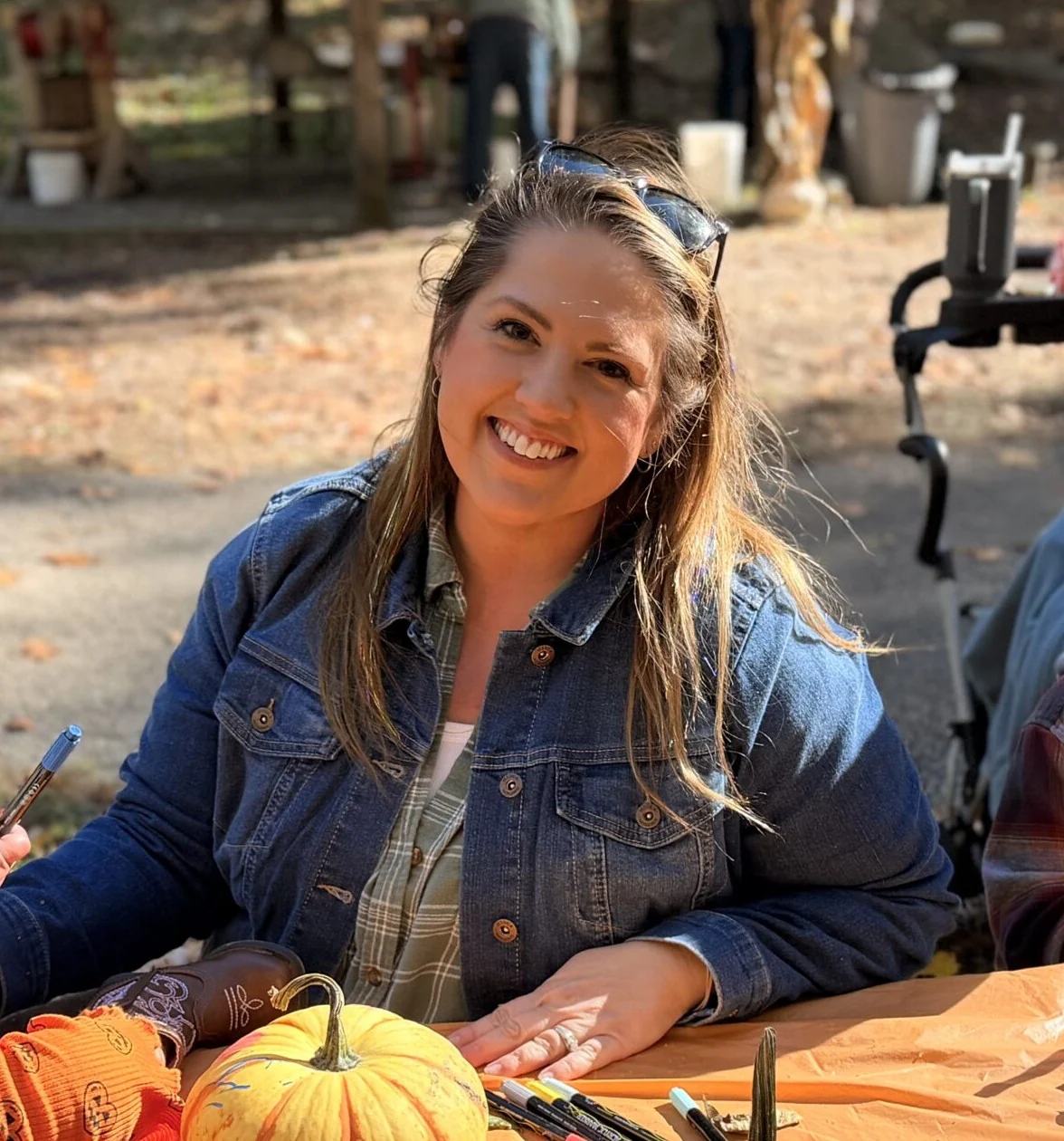 Jenn smiling at an outdoor craft table in autumn, in a denim jacket over a plaid shirt, sunglasses on her head