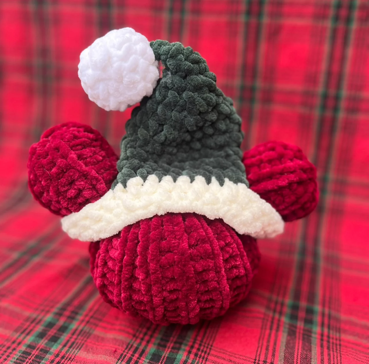 A deep red crocheted Magic Mouse pumpkin wearing a green Santa hat with a white pom-pom, on red plaid