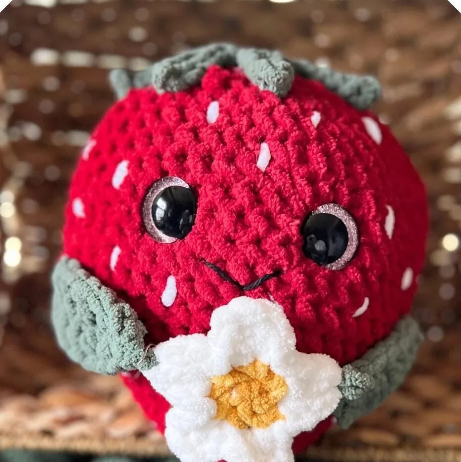 A red crocheted strawberry with safety eyes, a stitched smile, green leaf arms and a white flower with a yellow centre